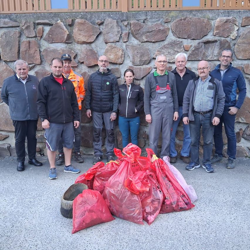 Eine Gruppe von Menschen in Jacken steht vor einer Steinmauer. Sie halten rote Plastiktüten. Ein Mann trägt einen Helm.