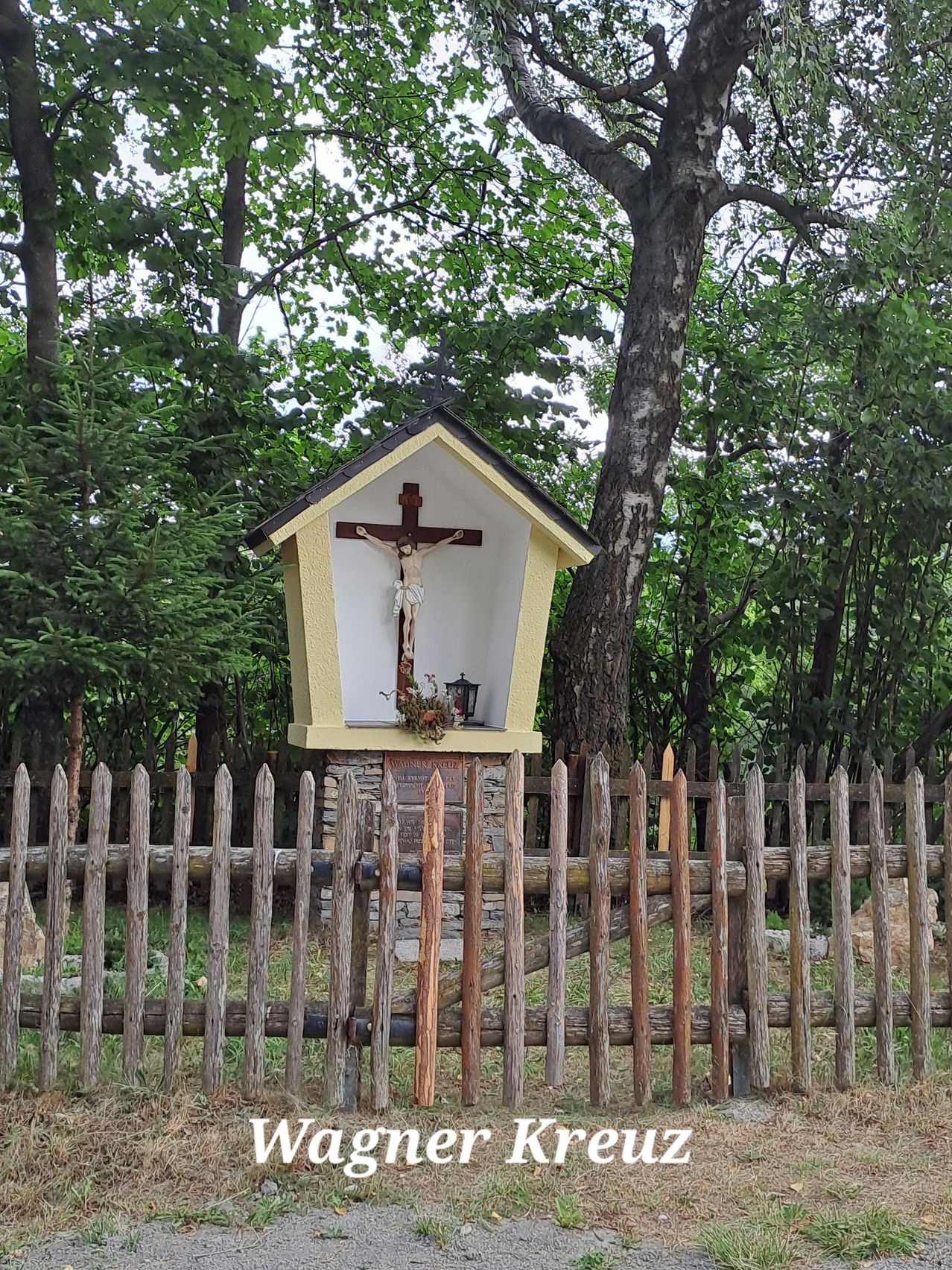 A small wooden chapel with a crucifix and a lamp inside, surrounded by a wooden fence and trees.