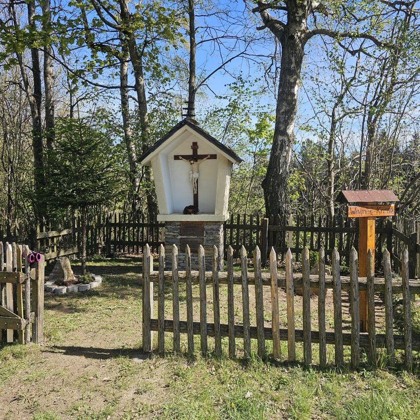 A small wooden chapel with a cross on top is surrounded by a wooden fence in a grassy area.