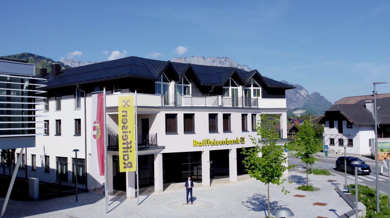 Ein modernes Gebäude mit schwarzen Dächern und Balkonen. Es zeigt ein gelbes Banner mit dem Wort 'Bauleitplanung' und einen Mann davor. Vor dem Gebäude steht ein Baum.