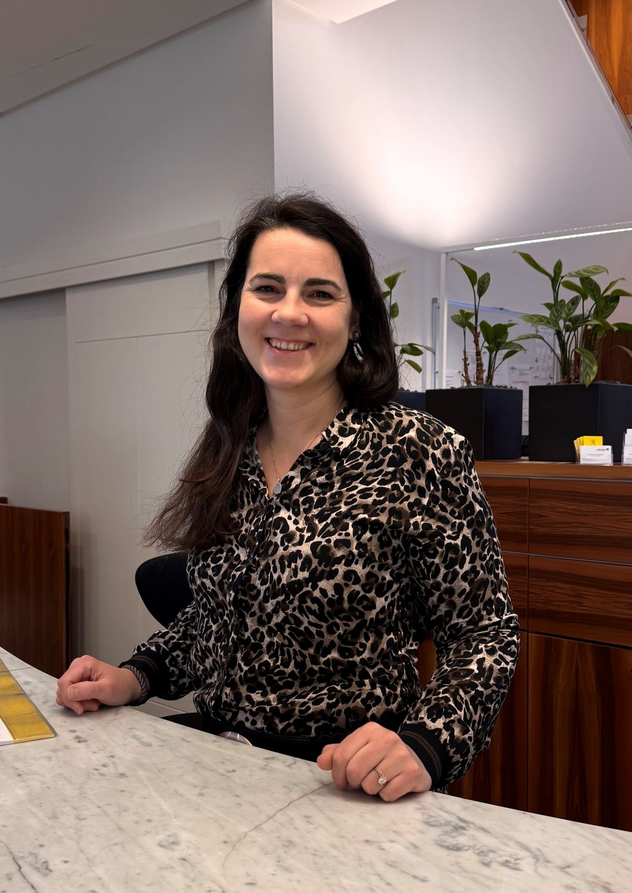 Eine lächelnde Frau in einem Leopardenmusterhemd sitzt an einem Schreibtisch in einem Büro. Hinter ihr befinden sich Holzschränke und eine weiße Wand mit einem Spiegel. Drei Topfpflanzen stehen auf dem Schrank.