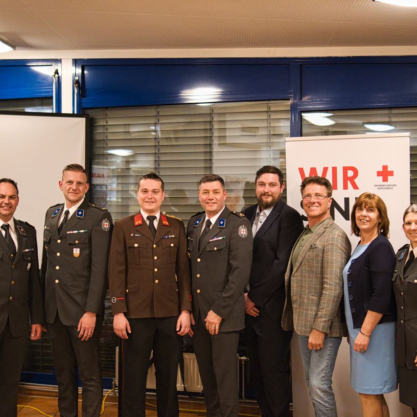 Mehrere Männer und Frauen in Uniformen und Anzügen posieren für ein Foto vor einem Schild, das Wir + N sagt.