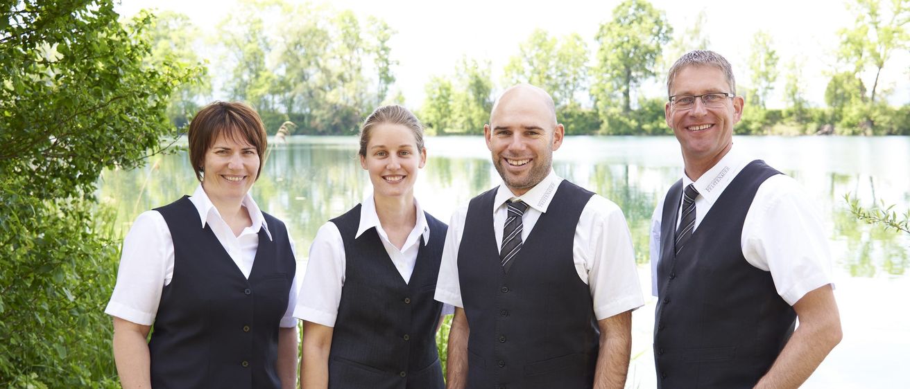 Vier Personen, zwei Frauen und zwei Männer, stehen nebeneinander vor einem See. Sie tragen alle passende schwarze Westen und weiße Hemden.