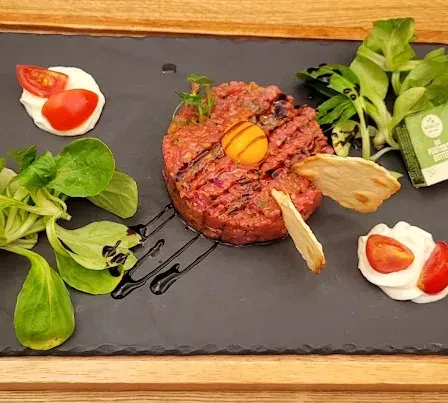 A gourmet dish with a red meat patty, topped with a fried egg, accompanied by green leaves, sliced tomatoes, and a dollop of cream on a black slate plate.