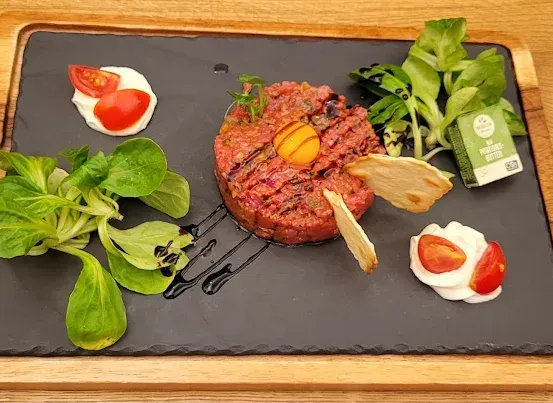 Ein Gourmetgericht mit einem roten Fleischpatty, gekrönt von einem gebratenen Ei, begleitet von grünen Blättern, geschnittenen Tomaten und einem Klecks Sahne auf einem schwarzen Schieferteller.