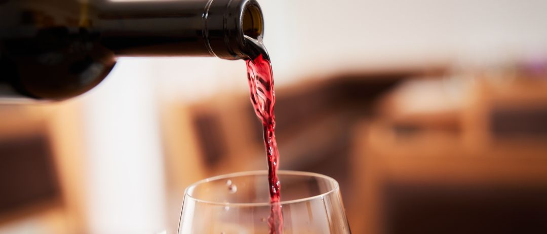 Eine Flasche Rotwein wird in ein Weinglas gegossen. Der Wein fließt in einem dünnen Strahl herunter. Der Hintergrund mit hölzern Möbeln ist unscharf.