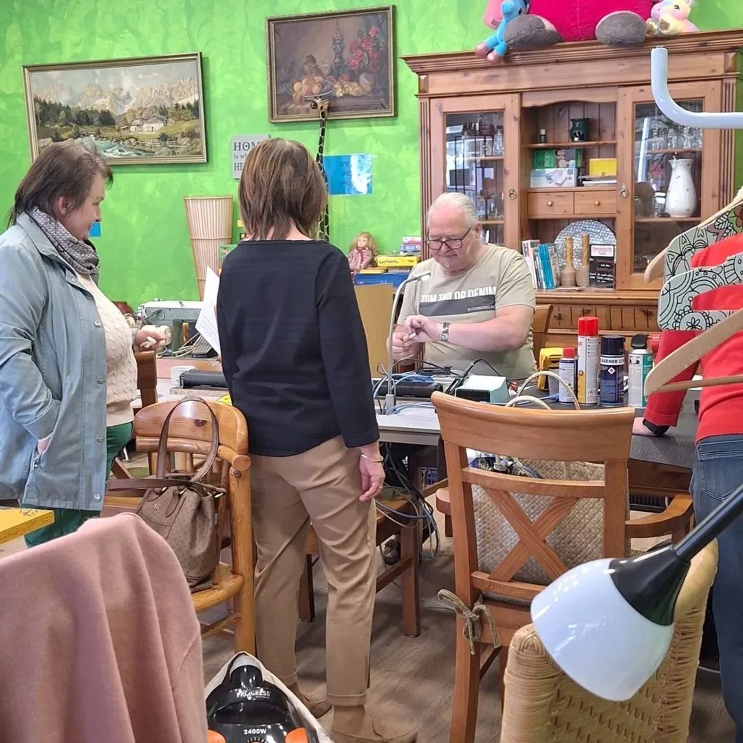 Ein älterer Mann arbeitet an einem Gerät, während er an einem Tisch im Esszimmer sitzt. Mehrere Personen stehen in der Nähe, darunter eine Frau in einem Schal.