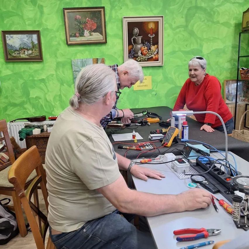 Drei Personen arbeiten in einem Raum mit grünen Wänden an einem Projekt. Ein Mann sitzt mit Werkzeugen am Schreibtisch, ein anderer Mann lehnt sich über ihn und eine Frau steht hinter ihm.