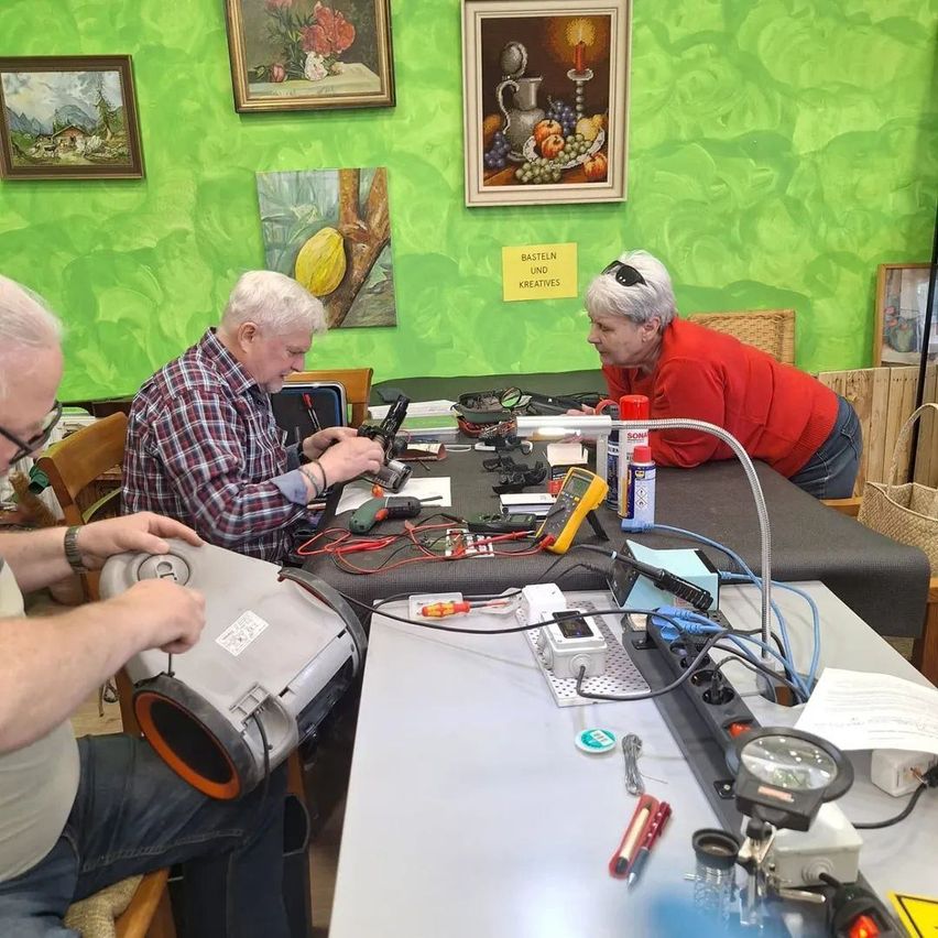 Drei Personen sitzen um einen Tisch mit verschiedenen Werkzeugen. Ein Mann repariert ein Gerät, eine Frau lehnt sich über ihn und der dritte Mann hält ein Gerät. Der Tisch ist mit Werkzeugen und Kabeln übersät. Hinter ihnen ist eine grüne Wand mit gerahmten Bildern zu sehen.