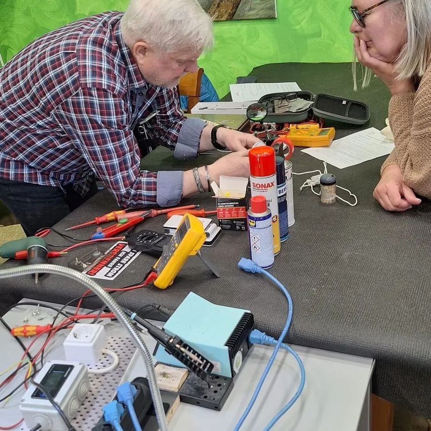 Ein älterer Mann und eine Frau arbeiten an einem Projekt an einem Tisch mit verschiedenen Werkzeugen und Materialien.