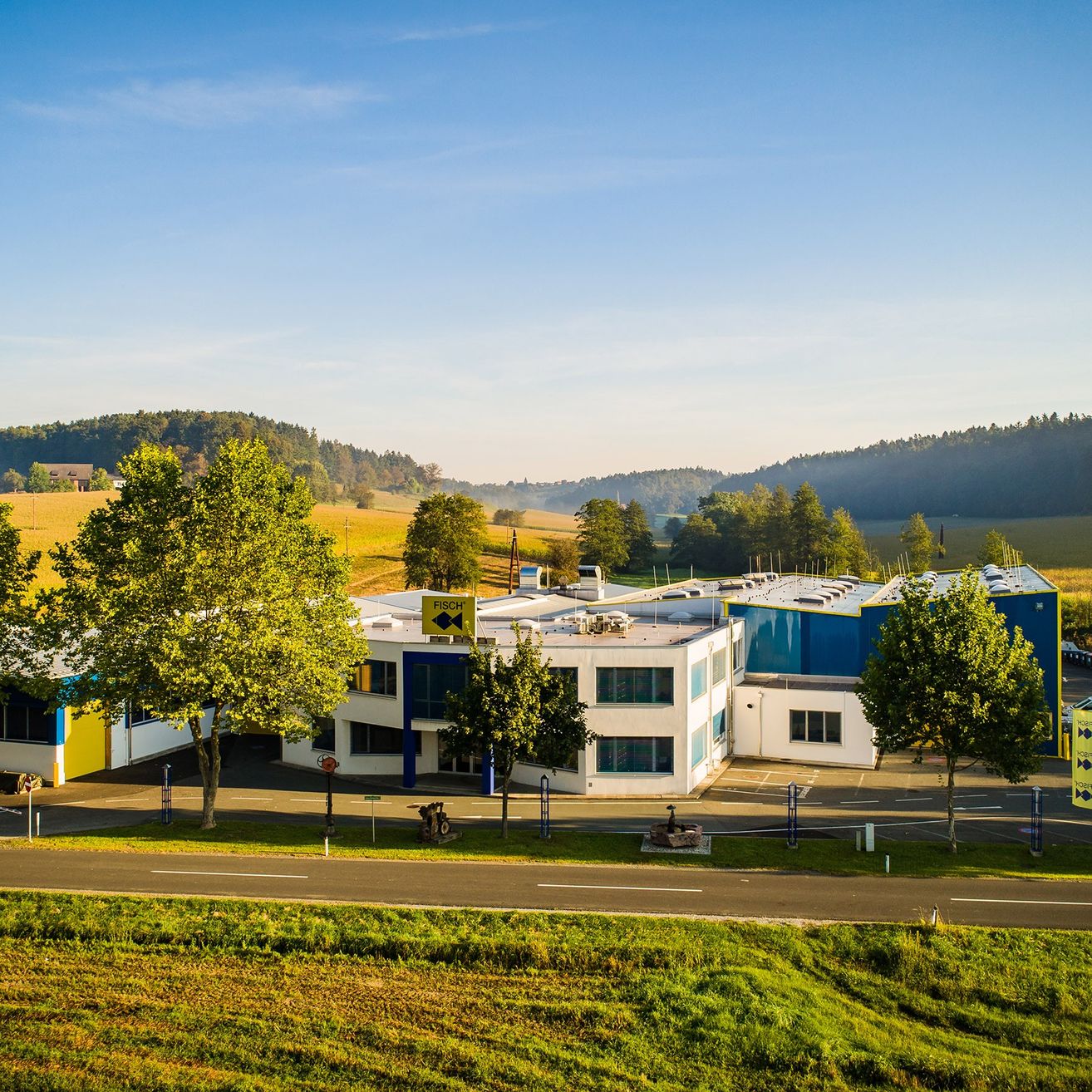 Eine Luftaufnahme eines Gebäudes mit blauen und weißen Wänden, umgeben von Gras, Bäumen und Bergen unter einem klaren blauen Himmel.