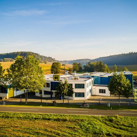 Eine Luftaufnahme eines Gebäudes mit blauen und weißen Wänden, umgeben von Gras, Bäumen und Bergen unter einem klaren blauen Himmel.