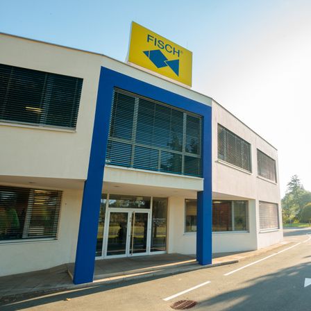 Ein Gebäude mit einem gelben Schild, das 'Fisch' liest, hat blaue Akzente und einen Glaseingang. Es befindet sich auf einer Straße mit Bäumen an der Seite.