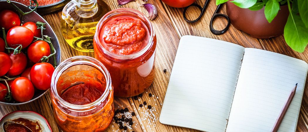 Ein Holztisch mit zwei Gläsern roter Sauce, einem Tomatenteller, einer Ölflasche, einer Knoblauchzehe und einem offenen Notizbuch.