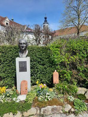 Eine Büste eines Mannes steht in einem Garten mit einer Plakette, umgeben von einer Vielzahl von Blumen, Büschen und Steinen. Ein schlossartiges Gebäude ist im Hintergrund sichtbar.