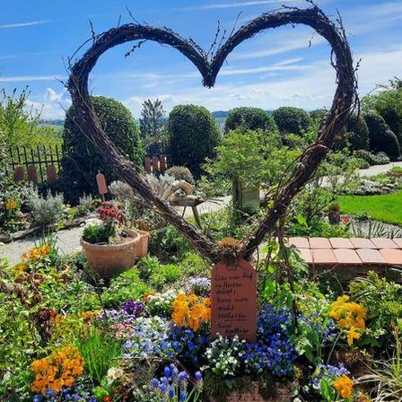 Ein herzförmiges Gebilde aus Zweigen steht in einem Garten mit verschiedenen Blumen und Pflanzen. Eine Tafel ist in der Mitte des Herzens platziert. Der Garten ist von einem Zaun umgeben, und ein Weg führt zum Herzen. Der Himmel ist blau mit einigen Wolken.