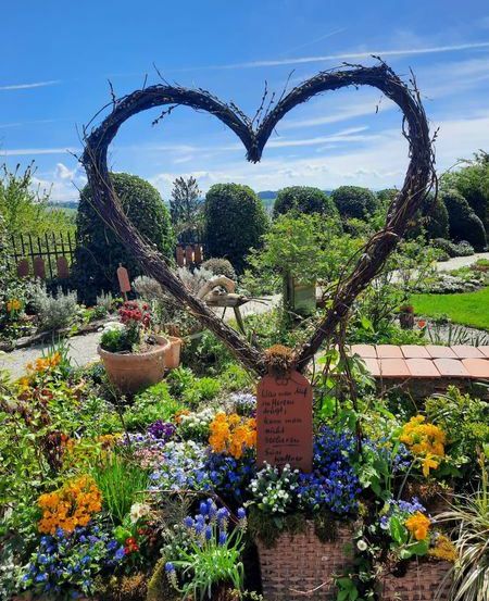 Ein herzförmiges Gebilde aus Zweigen steht in einem Garten mit verschiedenen Blumen und Pflanzen. Eine Tafel ist in der Mitte des Herzens platziert. Der Garten ist von einem Zaun umgeben, und ein Weg führt zum Herzen. Der Himmel ist blau mit einigen Wolken.