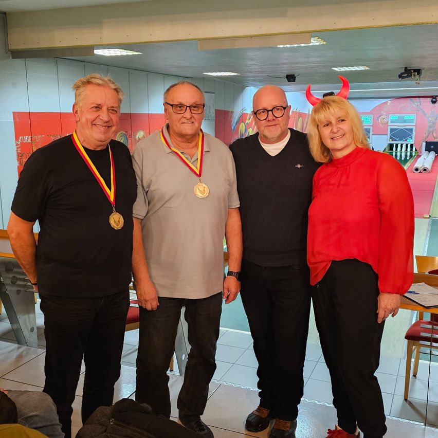 Vier Menschen stehen zusammen in einem Raum, drei Männer und eine Frau. Die Männer tragen Medaillen um den Hals. Sie lächeln und scheinen für ein Foto zu posieren. Hinter ihnen befindet sich eine rote Wand mit verschiedenen Objekten.