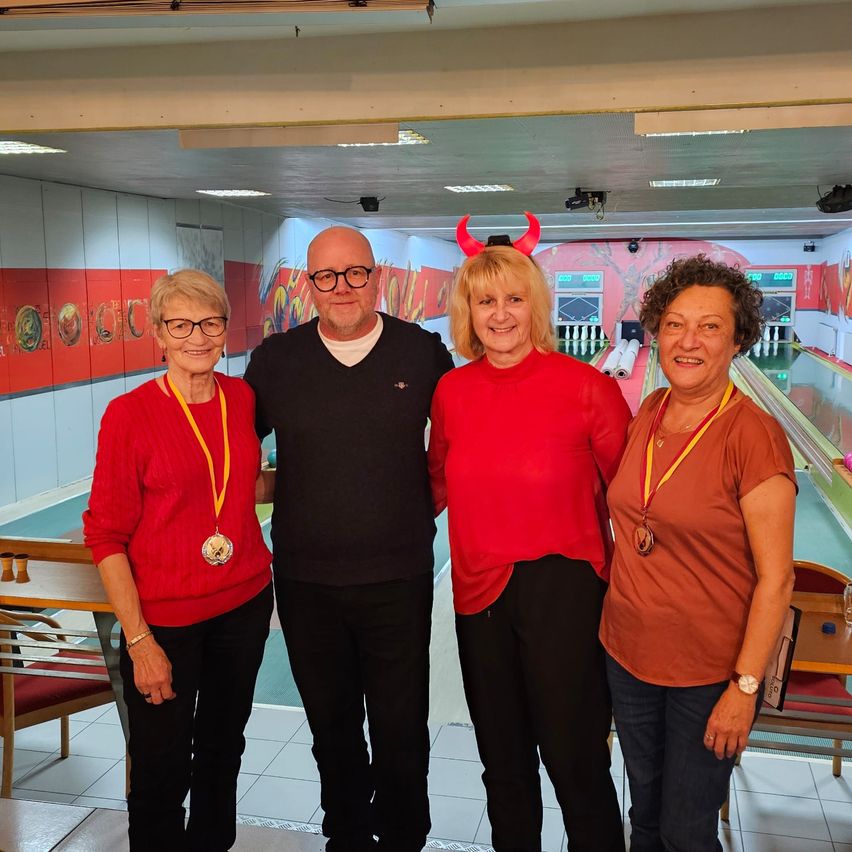 Fünf Personen stehen in einer Bowlingbahn. Vier Frauen in roten und braunen Shirts und ein Mann in einem schwarzen Shirt. Alle lächeln und tragen Medaillen. Die Bowlingbahn hat farbenfrohe Wände.