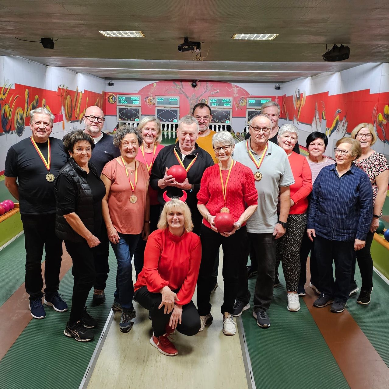 Eine Gruppe Erwachsener in einer Bowlingbahn, posiert für ein Foto, einige halten Bowlingkugeln und tragen Medaillen. Die Bowlingbahn hat farbenfrohe Wände mit Bowlingkegeln und digitalen Anzeigetafeln.