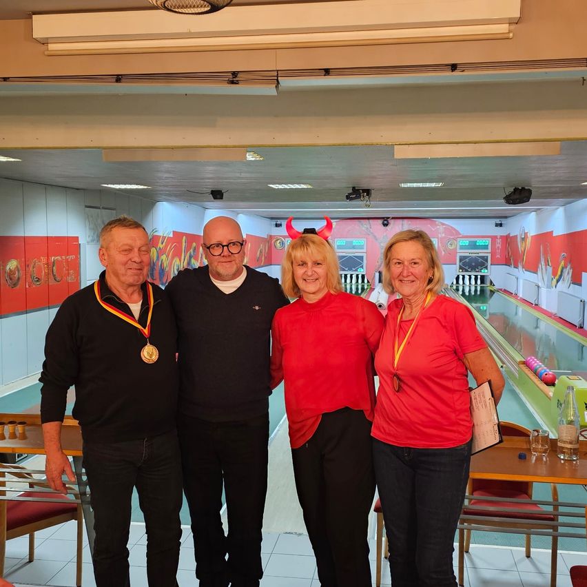 Vier Personen stehen in einer Bowlingbahn. Drei tragen rote Hemden, ein Mann eine Medaille. Sie lächeln für die Kamera.
