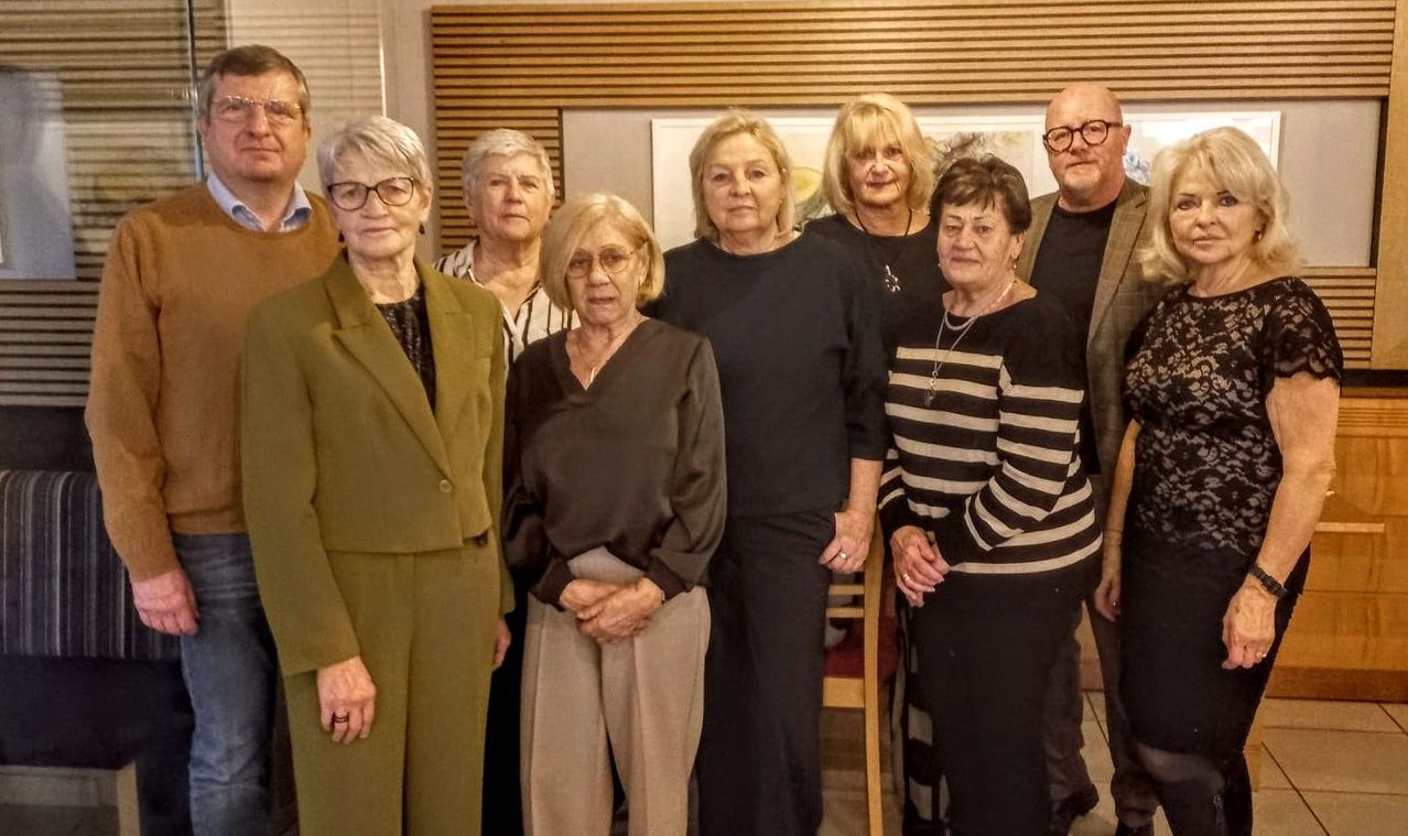 Eine Gruppe älterer Personen steht in einem Raum zusammen, lächelt und posiert für ein Foto. Sie sind in verschiedene Winterkleidung gekleidet, und es gibt zwei Gemälde an der Wand hinter ihnen.