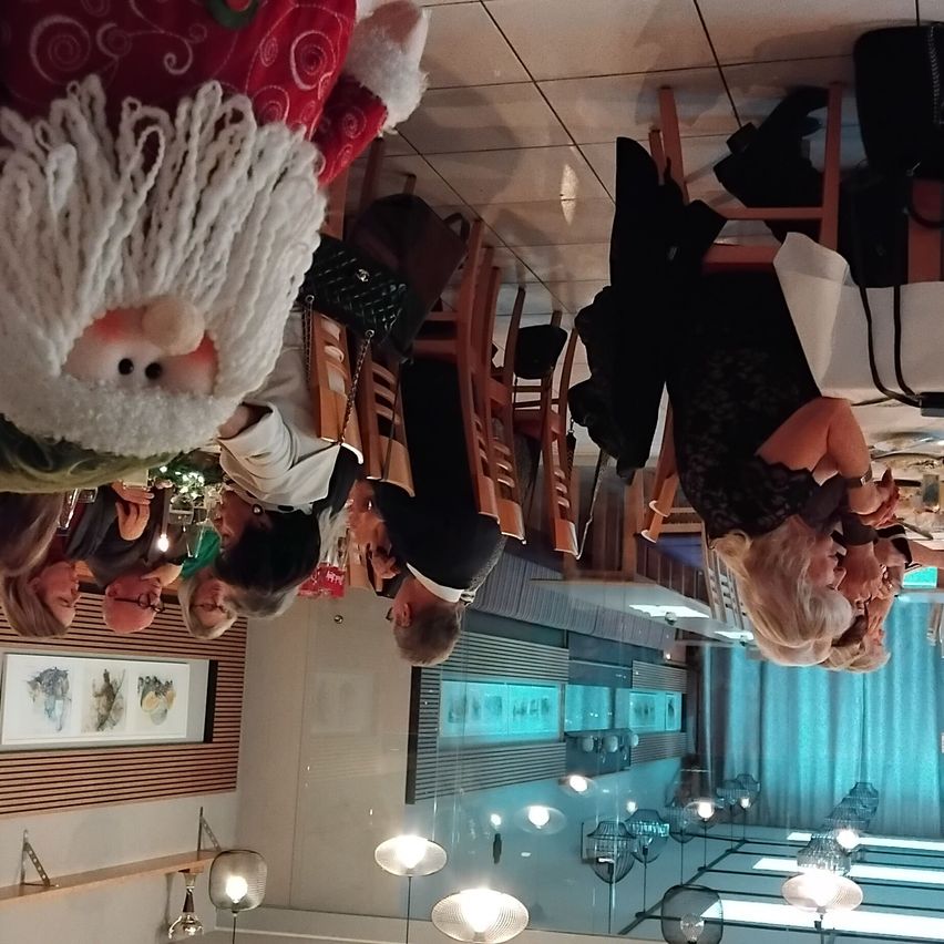 Eine Gruppe von Menschen wird von oben gesehen, versammelt in einem Restaurant. Eine Person, die eine Weihnachtsmann-Kostüm trägt, ist in der Mitte. Mehrere Stühle und Tische sind angeordnet, und Jacken hängen an den Stühlen. Die Decke hat Lampen und gerahmte Bilder.