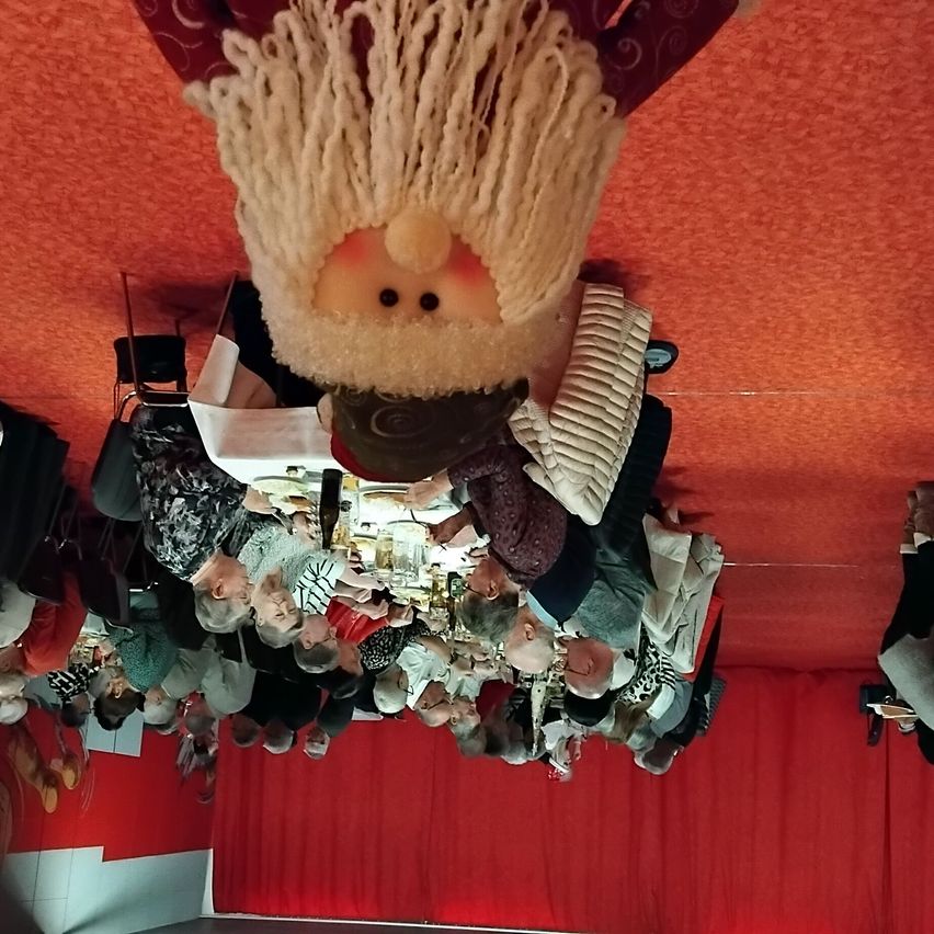 Eine Gruppe von Menschen sitzt um einen Tisch mit Getränken und Essen, mit einer puppenartigen Figur darüber. Der Raum hat rote Wände und einen roten Vorhang.