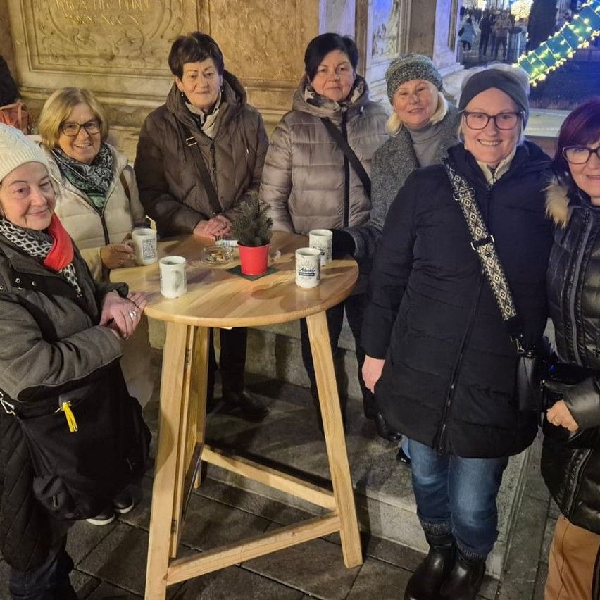 Eine Gruppe von Frauen in Winterkleidung steht lächelnd um einen kleinen runden Holztisch mit Tassen und einem Topf. Sie scheinen sich bei einer Veranstaltung im Freien zu befinden.