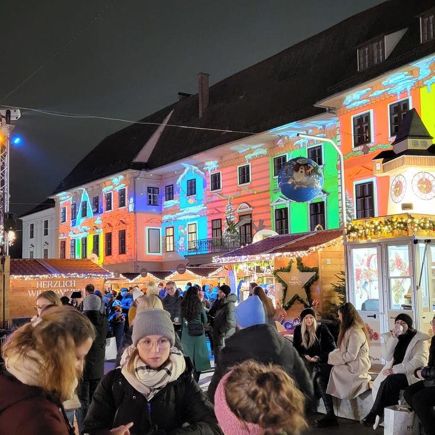 Bei Nacht versammelt sich eine Menschenmenge vor einem farbenfroh beleuchteten Gebäude mit festlicher Atmosphäre, einige sitzen, andere stehen. In der Nähe gibt es dekorierte Stände, an denen die Leute stöbern.