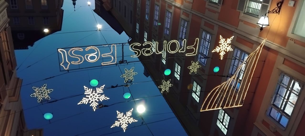 Ein Gebäude mit mehreren Fenstern spiegelt sich im Fluss wider. Die Lichter auf der Brücke lesen 'Frohe Weihnachten'. Schneeflocken hängen von der Brücke.