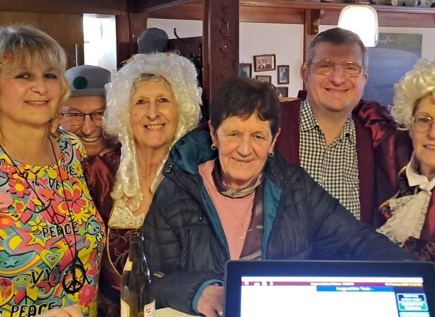 A group of adults are smiling and posing for a photo. They are wearing different clothing and accessories. A woman in a white wig and a man in glasses are sitting next to each other. A bottle is placed in front of them.