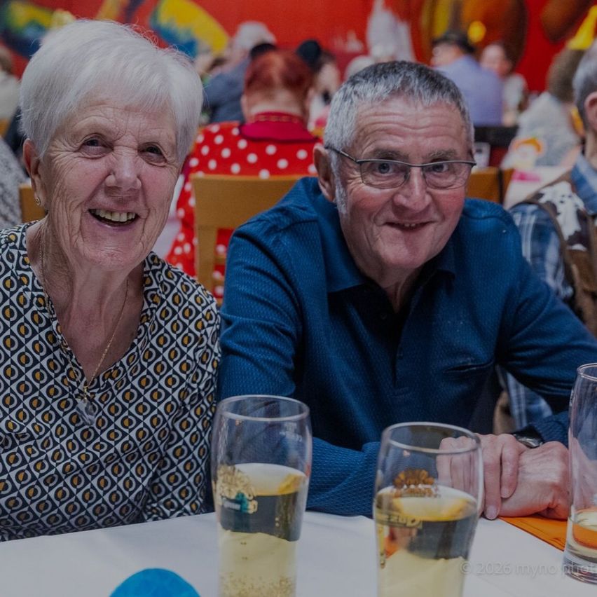 An elderly couple, both smiling, seated at a table with two glasses of beer, surrounded by a lively restaurant setting.