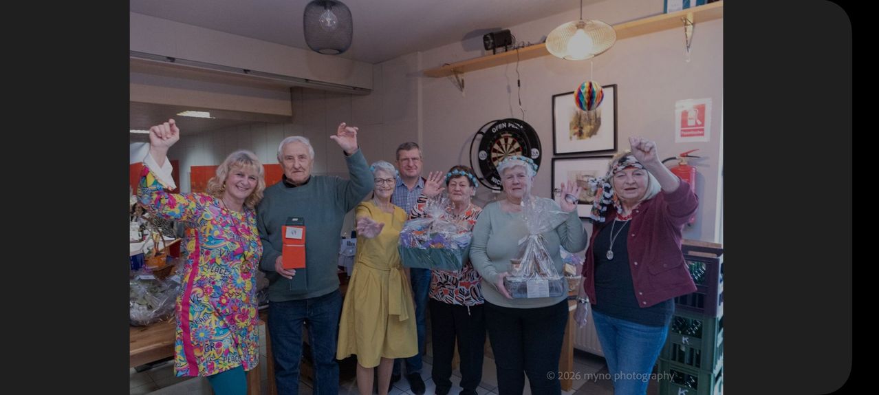 A group of people stand in a room, some holding gift baskets. A man raises his hand while a woman smiles. There are framed pictures on the wall and a dartboard. The image has a watermark.