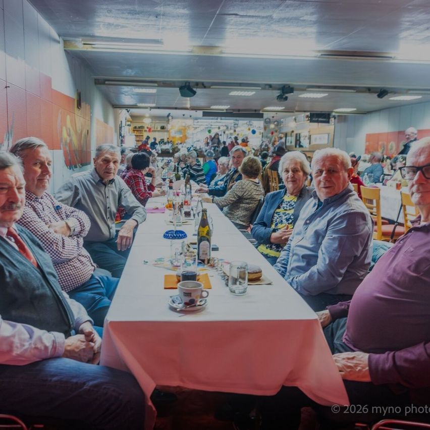 A group of older people are sitting at a long table in a restaurant. There are cups, plates, and bottles on the table. Some of them are looking at each other. The room has a white ceiling with lights.