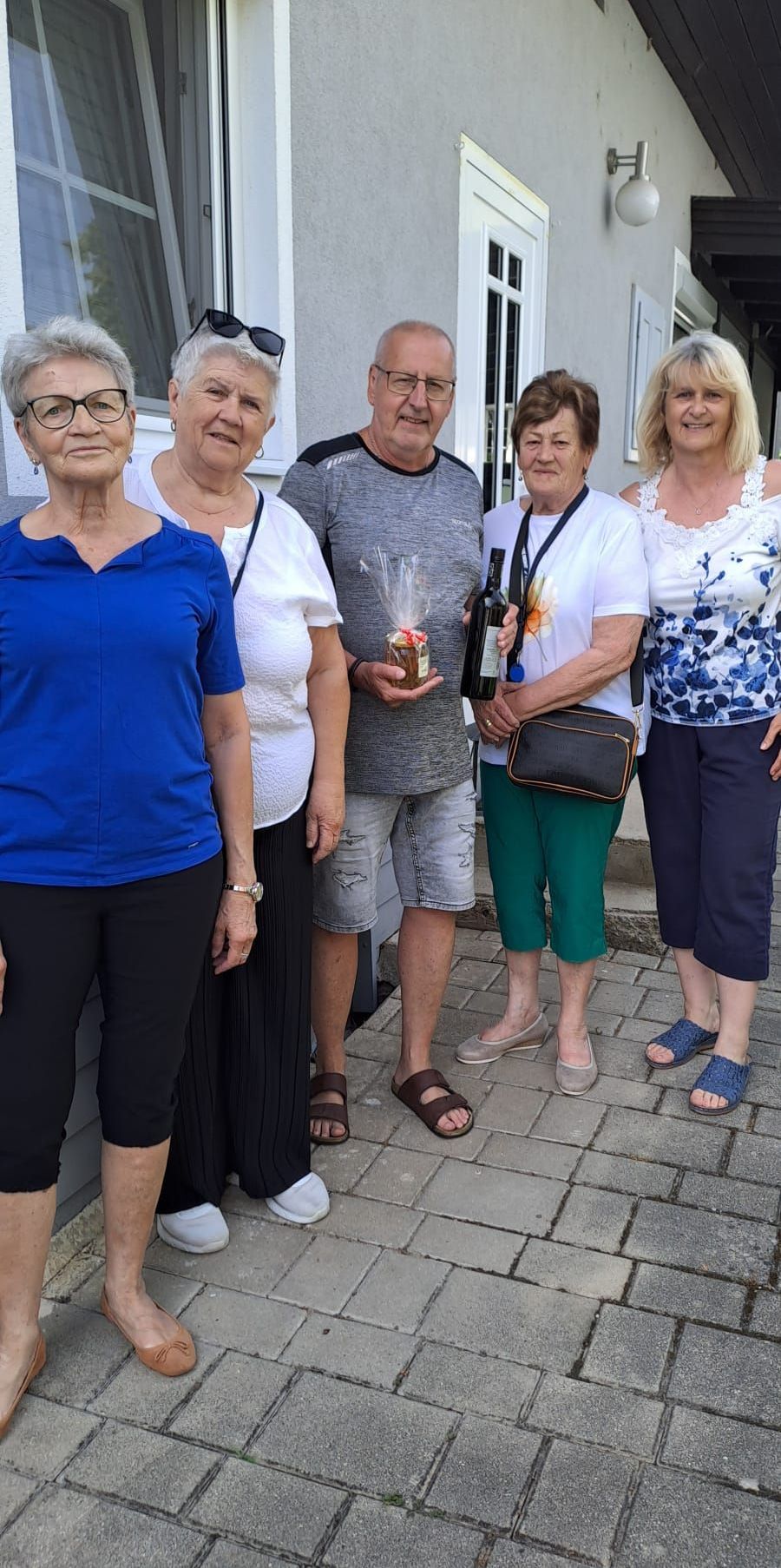 Five elderly individuals standing on a brick path. A man holds a bottle of wine and a small gift. Two women wear sleeveless tops, one has a handbag.