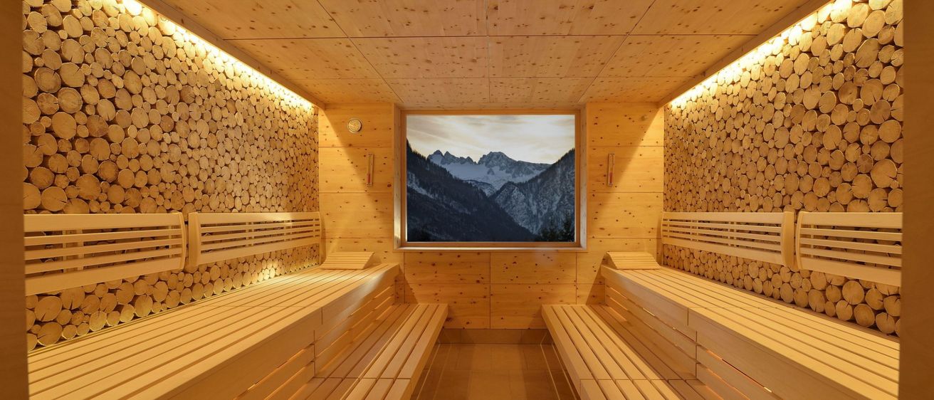 Eine Holzsauna mit Holzbänken und Wänden. Eine große Leinwand in der Mitte des Raumes zeigt eine Berglandschaft.