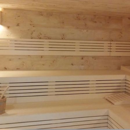 Innenansicht einer Sauna mit Holzbänken, einer wandmontierten Lampe und einem Holzeimer mit Schöpflöffel.
