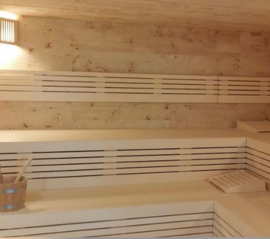 Innenansicht einer Sauna mit Holzbänken, einer wandmontierten Lampe und einem Holzeimer mit Schöpflöffel.
