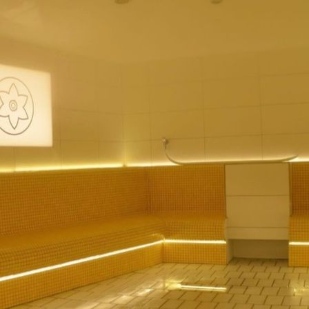 Ein Innenraum einer Sauna mit gelben gefliesten Wänden, einem weißen gefliesten Boden und einem quadratischen Fenster mit einem Blumenlogo. Es gibt zwei Bänke auf beiden Seiten, an der Wand ist ein Duschkopf befestigt.