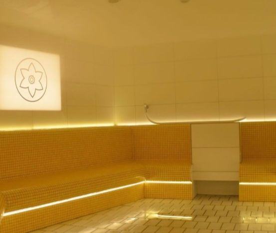 Ein Innenraum einer Sauna mit gelben gefliesten Wänden, einem weißen gefliesten Boden und einem quadratischen Fenster mit einem Blumenlogo. Es gibt zwei Bänke auf beiden Seiten, an der Wand ist ein Duschkopf befestigt.