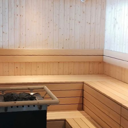 Innenraum einer Sauna mit Holzbänken und einem Heizgerät auf einem Ständer in der Ecke.