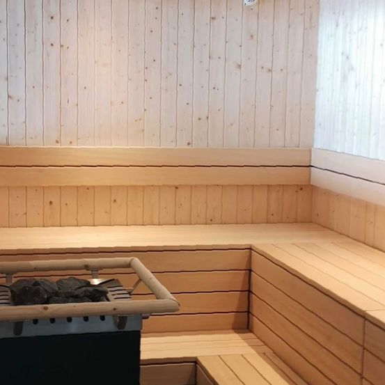 Innenraum einer Sauna mit Holzbänken und einem Heizgerät auf einem Ständer in der Ecke.