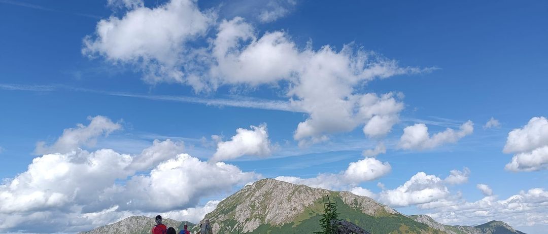 Eine Gruppe von Wanderern mit Rucksäcken steht auf einem Bergpfad unter einem klaren blauen Himmel mit Wolken. Die Bergkette ist im Hintergrund.