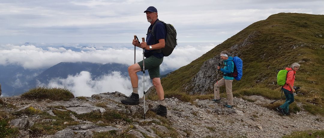 Zwei Wanderer mit Rucksäcken klettern mit Trekkingstöcken einen felsigen Bergpfad hinauf, unter einem bewölkten Himmel.