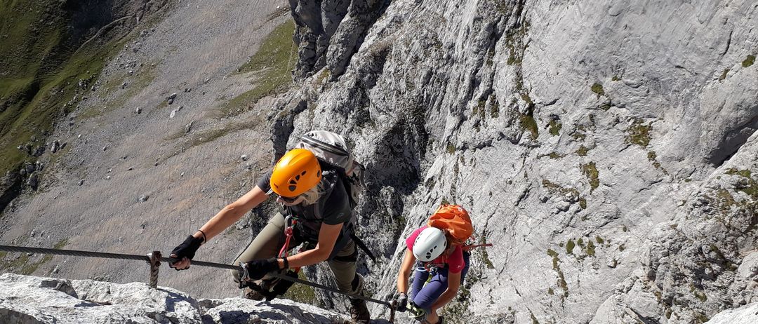 Zwei Kletterer in Helmen steigen einen steilen felsigen Berg hinauf, einer mit einem orangefarbenen Rucksack. Sie halten sich an einem Seil fest.