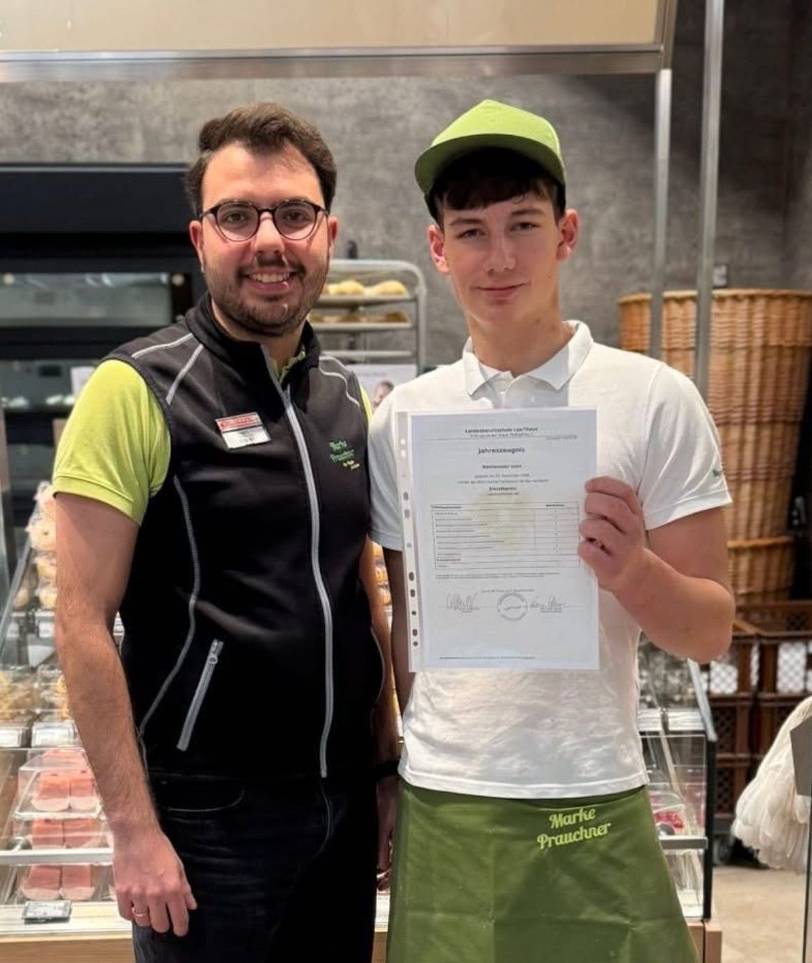 Zwei Männer posieren für ein Foto, einer hält eine Urkunde. Sie stehen in einer Bäckerei. Dahinter ist eine Vitrine mit Brot zu sehen.