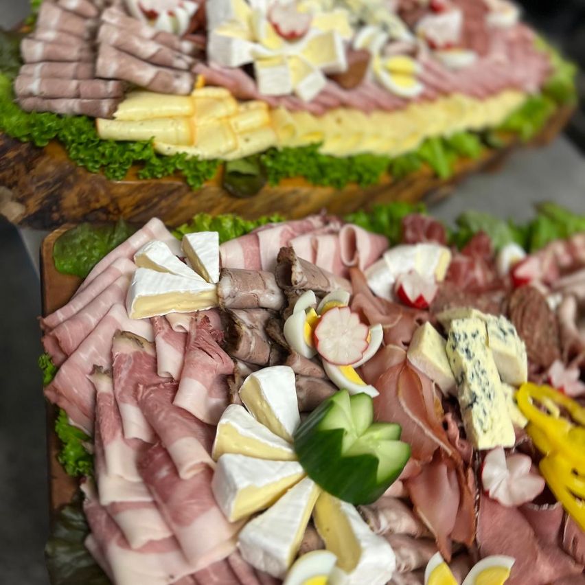 Zwei große Charcuterie-Bretter, eines im Vordergrund und eines im Hintergrund, gefüllt mit einer Vielzahl von Fleisch, Käse und Gemüse. Das Vordergrund-Brett hat geschnittene Fleischsorten, Käse und Gemüse wie Gurken und Radieschen. Das Hintergrundbrett ist mit Salat bedeckt und hat ähnliche Zutaten.