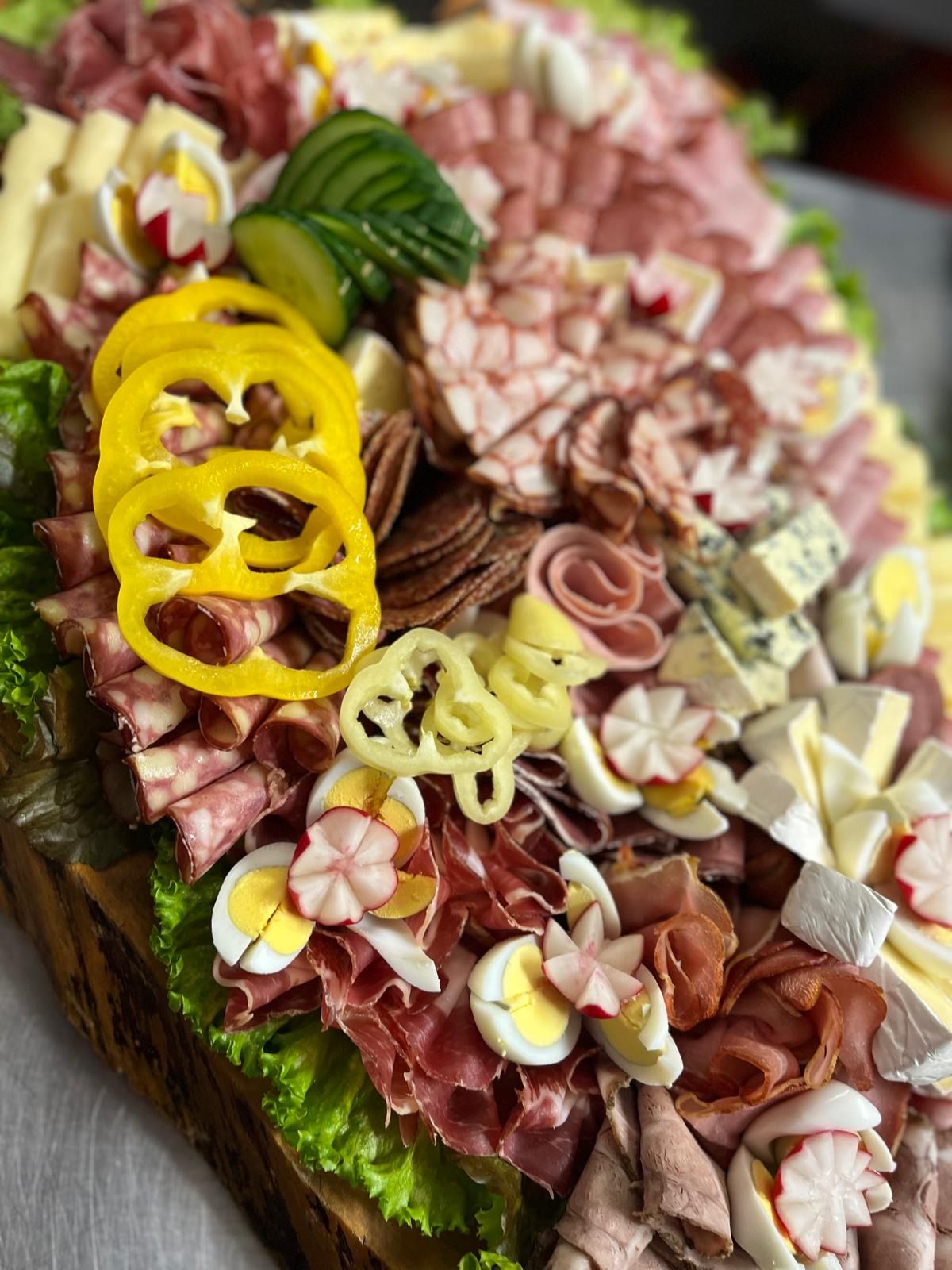 Eine Nahaufnahme einer Charcuterie-Platte mit geschnittenen Fleischsorten, Käse, Eiern und geschnittenem Gemüse. Es gibt geschnittene gelbe Paprika, geschnittene Gurken und Eier, die in Blütenformen angeordnet sind. Die Platte hat eine rustikale Holzbasis.
