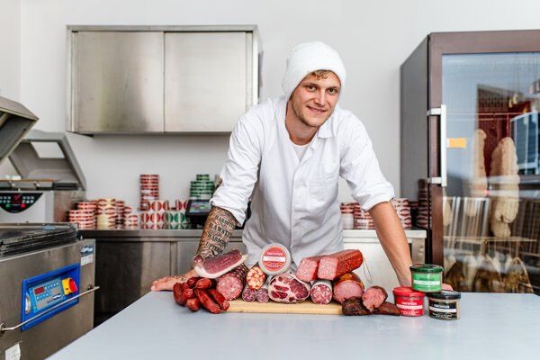 Ein Metzger posiert in einem Laden, umgeben von Fleisch auf einem Tresen und Kühlschränken im Hintergrund.